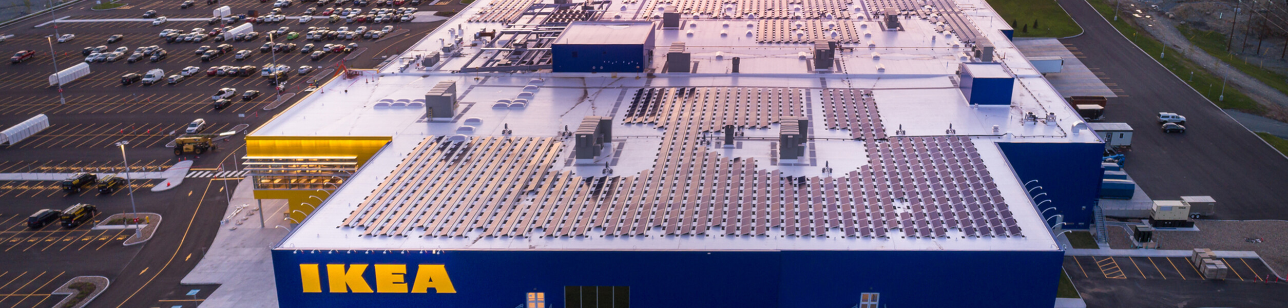 ikea-solaire Vue aérienne d'un grand magasin IKEA bleu avec des panneaux solaires sur le toit, entouré d'un grand parking et de routes. La scène est capturée au crépuscule, avec des lumières visibles à l'intérieur du bâtiment.