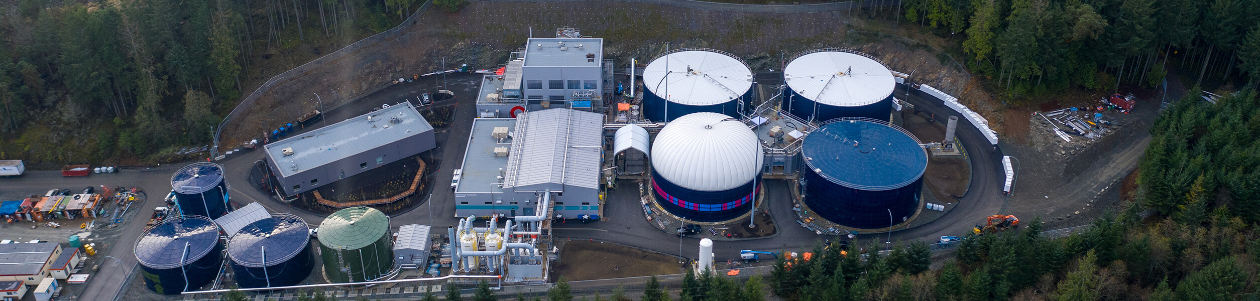 crd Vue aérienne d'une installation industrielle de traitement des eaux entourée d'une forêt dense, avec plusieurs grands réservoirs circulaires, des bâtiments rectangulaires et des véhicules de construction sur le site.