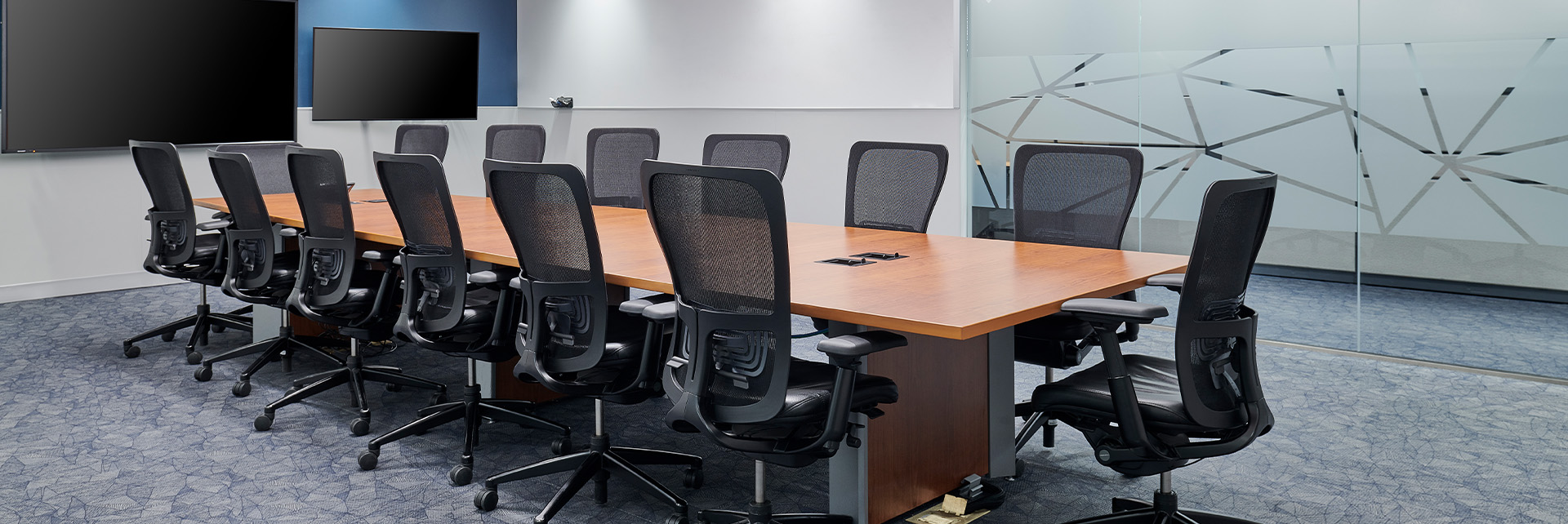 slider_tenant fit-ups A modern conference room with a long wooden table surrounded by black office chairs, two large wall-mounted screens, carpeted floor, and glass walls with geometric frosted patterns.