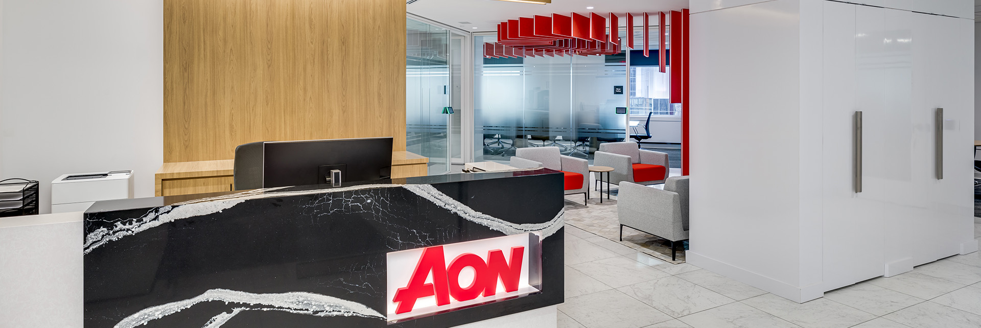 slider_renovations Modern office reception area with a black marble desk displaying a red Aon sign, light wood and white walls, glass partition, and gray chairs with red ceiling features in the background.