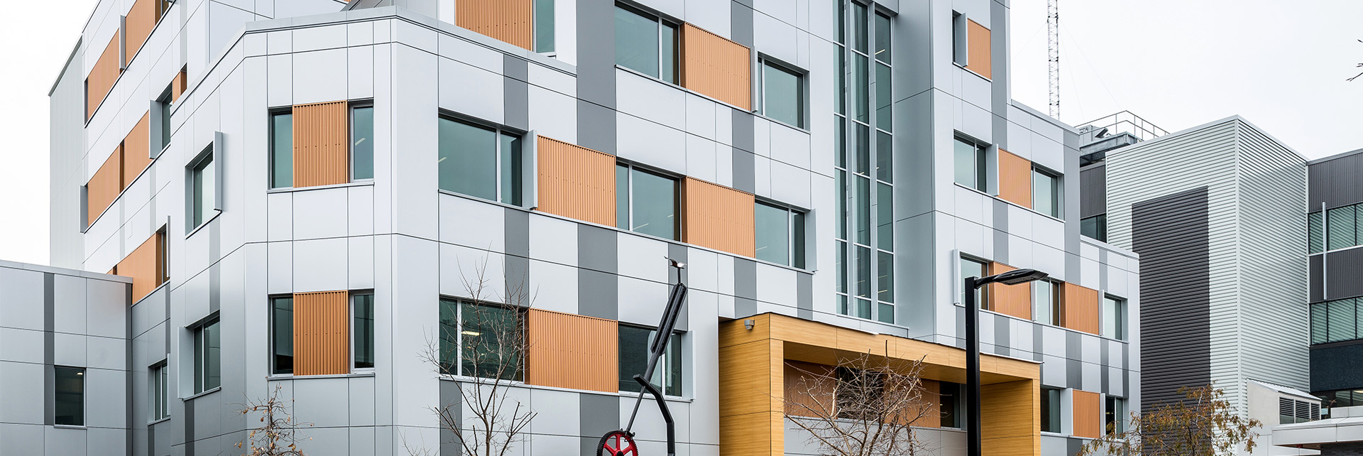slider_institutional A modern multi-story building with gray and orange panels, large windows, and a wooden entrance canopy. Some trees and a black street lamp are visible in front.