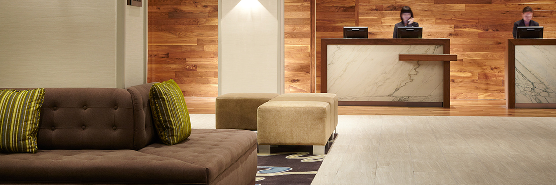 slider_hospitality Modern hotel lobby with a brown sofa, green-striped pillows, beige ottomans, and a rug. In the background, two receptionists stand behind marble and wood front desks. Wall features warm wood paneling.