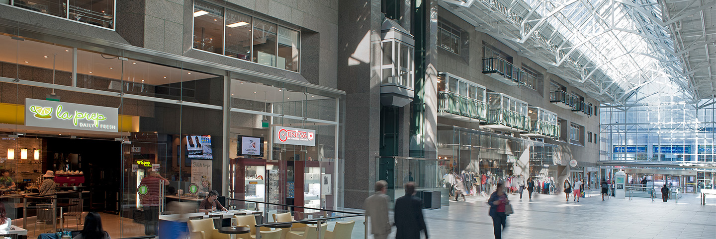 slider_comerciial A spacious, modern indoor atrium with high glass ceilings, restaurants, cafes, and shops along the sides. Several people walk or sit at tables, creating a lively, bustling atmosphere.