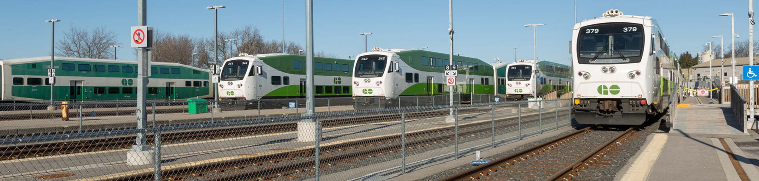 lincolnville-go-statione46f Une rangée de trains verts et blancs de GO Transit est garée dans une gare par temps clair, avec un train près du quai et plusieurs autres visibles derrière une clôture.