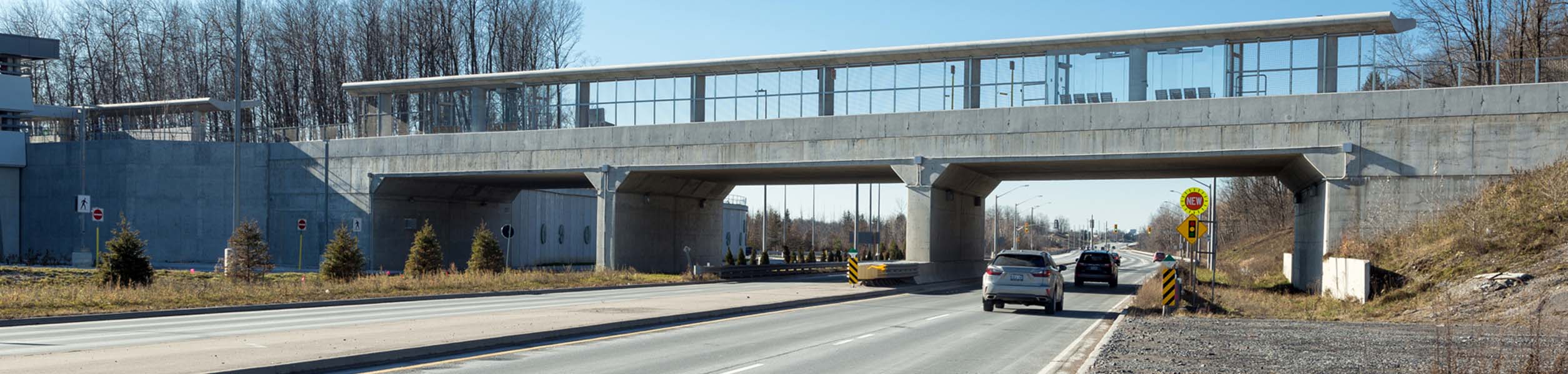 bloomington-rd-bridgeee1a Un viaduc en béton surmonté d'une structure moderne enjambe une route à chaussées séparées sous laquelle passent plusieurs voitures ; des arbres et des talus herbeux bordent les deux côtés sous un ciel dégagé.